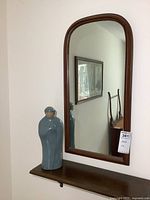 Framed wall mirror with a medium-tone wood frame and rounded top corners, 14 by 27 inches in size, resting above a narrow wooden shelf.