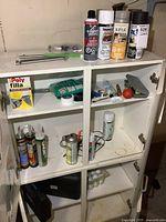 White cabinet with two shelves showing contents including several spray paint cans, caulking tubes, soldering kit, small socket kit and miscellaneous items.