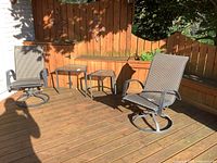 Pair of patio swivel chairs with metal frames and fabric seats on a wooden deck with two metal and glass side tables between them.