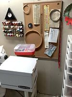Sewing accessories mounted on corkboard including wooden embroidery hoops, scissors, measuring ruler, and a container with thread bobbins, plus boxes and bins of sewing patterns below.
