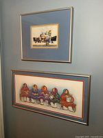 Two framed prints on blue wall; top print smaller with group of people and boat, bottom print larger with six women in traditional clothes