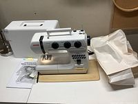 Sewing machine on table with hard case, dust cover, manual, and accessory pack.