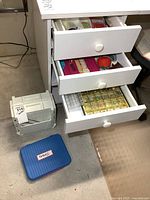 Wide view of white drawer unit with three drawers open showing various sewing notions and two additional thread boxes on the floor.