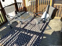 Outdoor patio set on wooden deck showing two metal mesh sling chairs, mesh round table, small stool and large geometric patterned outdoor carpet underneath.
