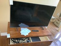 Front view of Samsung TV on wooden surface with remote, manuals, and part of HDMI cables visible in open drawer underneath.