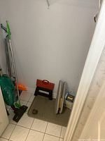 Full view of closet contents showing two Swiffers, green Swiffer in front with blue Swiffer behind; upright teal Miele vacuum; black step stool with red handle; orange and gray ironing board; gray ironing board cover; and iron with blue reservoir. White tiled floor and white walls.