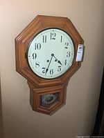 Front view of the vintage wooden wall clock with octagonal wooden frame and white clock face showing numerals 1 to 12, plus lower glass compartment.
