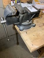 Left angled view of black 4-inch metal vise mounted on wooden workbench with tightening handle extended.