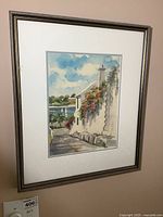 Framed watercolour painting showing coastal scene with building, flowers, walkway, water, sailboats, and sky.
