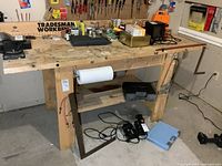 Wide view of heavy duty wooden workbench showing large solid wood top with tools and items present but not included in sale. Paper towel holder mounted below surface. Thick sturdy legs and support braces visible.