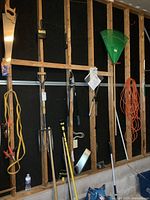 Wall mounted yard and automotive tools including pitchfork, shovel, rake, brush, extension cord, pole light changer, and bags of ice melter on the floor.