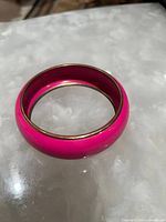 Photo of a single pink enamel bangle bracelet with gold-tone trim placed on a reflective surface.