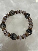 Full view of the beaded bracelet showing the combination of large grey decorative beads, purple beads and metallic spacers on a soft white fabric.