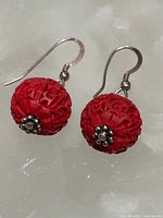 Close-up photo of a pair of red cinnabar-style carved spherical earrings with silver-colored metal hooks and decorative metal flower caps at the base of each bead.