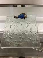 Square glass writing board with bubble texture and blue silver fish figurine from a top angle.