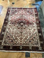 Full view of rectangular antique-looking wool rug with geometric floral patterns in earth tones on a light background lying on stone floor.