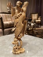 Angle showing the figure seated on a mythical bird with detailed carving on wings and tail, and the man holding a mask.