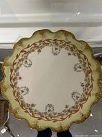 Top view of front of plate on reflective surface, showing scalloped shape, floral wreath design and gold trim