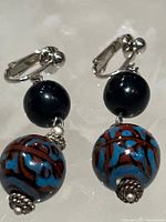 Pair of clip-on earrings each featuring a large blue bead with red abstract patterns and silver-tone metal embellishments. Shown against a beige background.