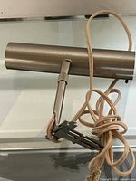Oil painting light with tubular metal hood, attached clamp, and beige power cord coiled beside it