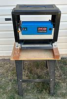 Front view of Ryobi 10" surface planer mounted on a black metal stand with wooden supports, showing overall condition and structure.