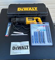 Full view of DEWALT DW303M reciprocating saw on top of the DEWALT carrying case with power cord wrapped, instruction manual, loose saw blades, and Mastercraft blade set on table.