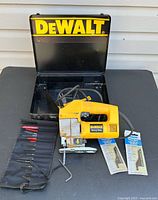 Jig saw with blade holder, extra blades and hex key in front of open DEWALT metal case on black surface against light wall.