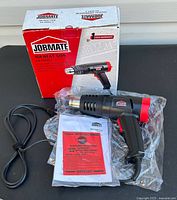 Heat gun in black and red with power cord and manual on black surface next to box.