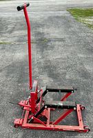 Photo showing red motorcycle jack with handle and black rubber lifting pads on pavement.