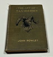 Front cover of The Art of Taxidermy book by John Rowley showing textured dark cover with gold lettering and ram head illustration.