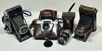 Four vintage cameras displayed side by side on white surface: Two folding bellows cameras, Voigtländer 35mm SLR in leather case, and additional lens with leather case