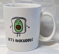 Front view of white ceramic mug with avocado in toast cartoon and 'Let's Avocuddle' text.