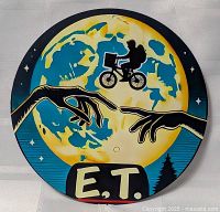 Front view of round tin sign depicting E.T. movie scene with silhouette of bike rider in front of moon and two hands reaching toward each other.