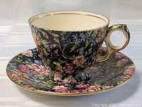 Tea cup and saucer set with floral pattern and gold trim on black background, cream interior of cup visible.