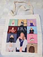 Front view of the white canvas tote bag showing colorful illustrations of Taylor Swift representing various eras with text in the center.