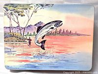 Top view of rectangular tin lid showing trout jumping out of water with mountainous landscape and fisherman in background in a colorful painted scene.