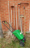 Wide shot of all long-handled tools leaning against brick wall with green spreader in front