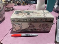 Closed view of ceramic rectangular trinket box with painted bird on lid and floral design on sides, placed next to a marker for scale.