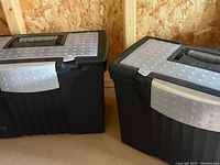 Two black plastic file folder totes with gray textured latching handles closed and placed side by side on a plywood floor in a wooden structure.