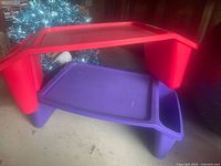 Photo showing two plastic lap desks, one red and one purple, stacked with visible side storage compartments and flat work surfaces.