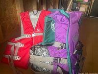 Two adult oversized life jackets, one red with gray straps and one purple with green accents and gray buckles, displayed hanging indoors.