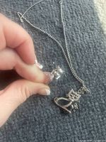 Close-up of a hand holding one sterling silver kitty earring showing detailed cat design with a gemstone eye, lying next to a matching kitty necklace with similar design and gemstone detail