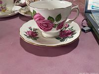 Side view of the bone china cup sitting on the saucer, showing the detailed pink rose floral design and gold trim.