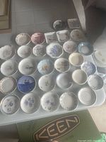 View of entire collection of approximately 33 assorted butter pat dishes arranged on table showing variety of designs and shapes.