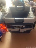 Photo of a large foldable fabric tote bag with wheels, black and gray in color, labeled 'Cutting Edge' on front. Multiple pockets and sturdy handles visible.