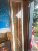 Tall and narrow IKEA Billy glass door in white, leaning against a wall with visible wood texture background and reflection on the glass panel.