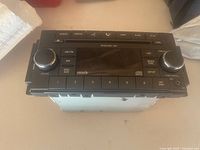 Front view of black car radio unit showing buttons, rotary knobs, and digital display with various control labels.