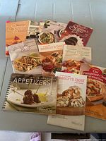 Photo showing 8 Pampered Chef cookbooks arranged on a table including multiple copies of Season's Best, Casual Impressive Appetizers, and Pampered Chef Favourites.