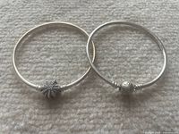 Two silver Pandora bracelets laid on a fabric surface. One bracelet has a floral clasp charm, the other a round charm engraved with 'PANDORA'.