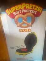 Photo of SuperPretzel Soft Pretzels Maker Machine with box showing prominently the pretzel image and text. The machine is yellow with a black heating plate inside and a red cheese melting pod.